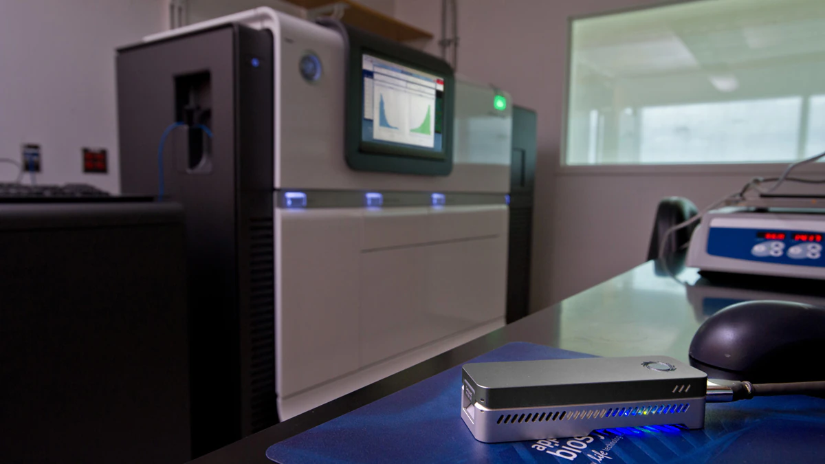 nanopore-sequencer-1200 A small nanopore sequencer (about the size of a short stapler) sits on a table in the foreground. In the background is a large sanger sequencer machine (the size of a large copy machine)