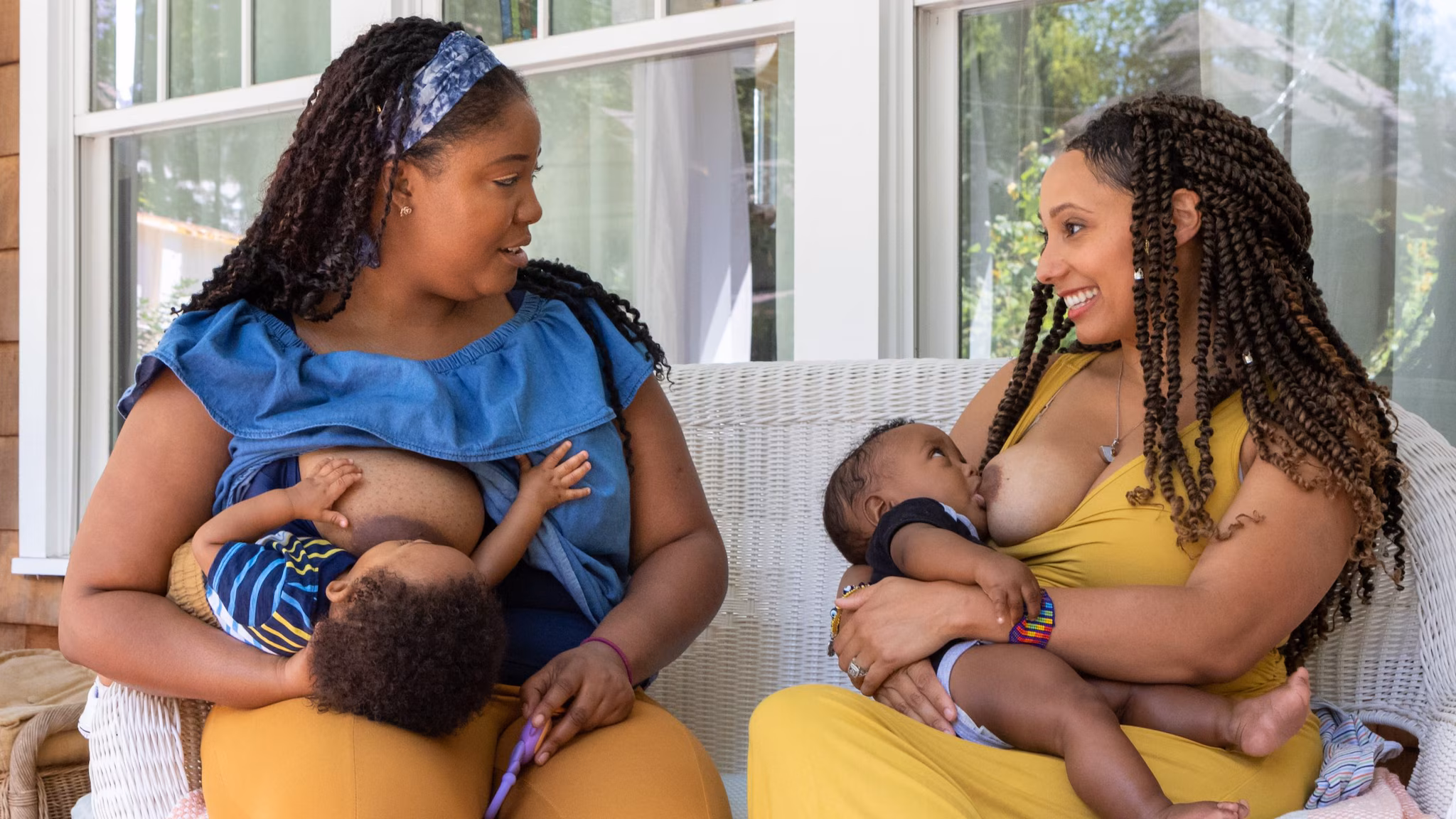 — title missing — Two women breastfeed two children while sitting on a porch outside a house.