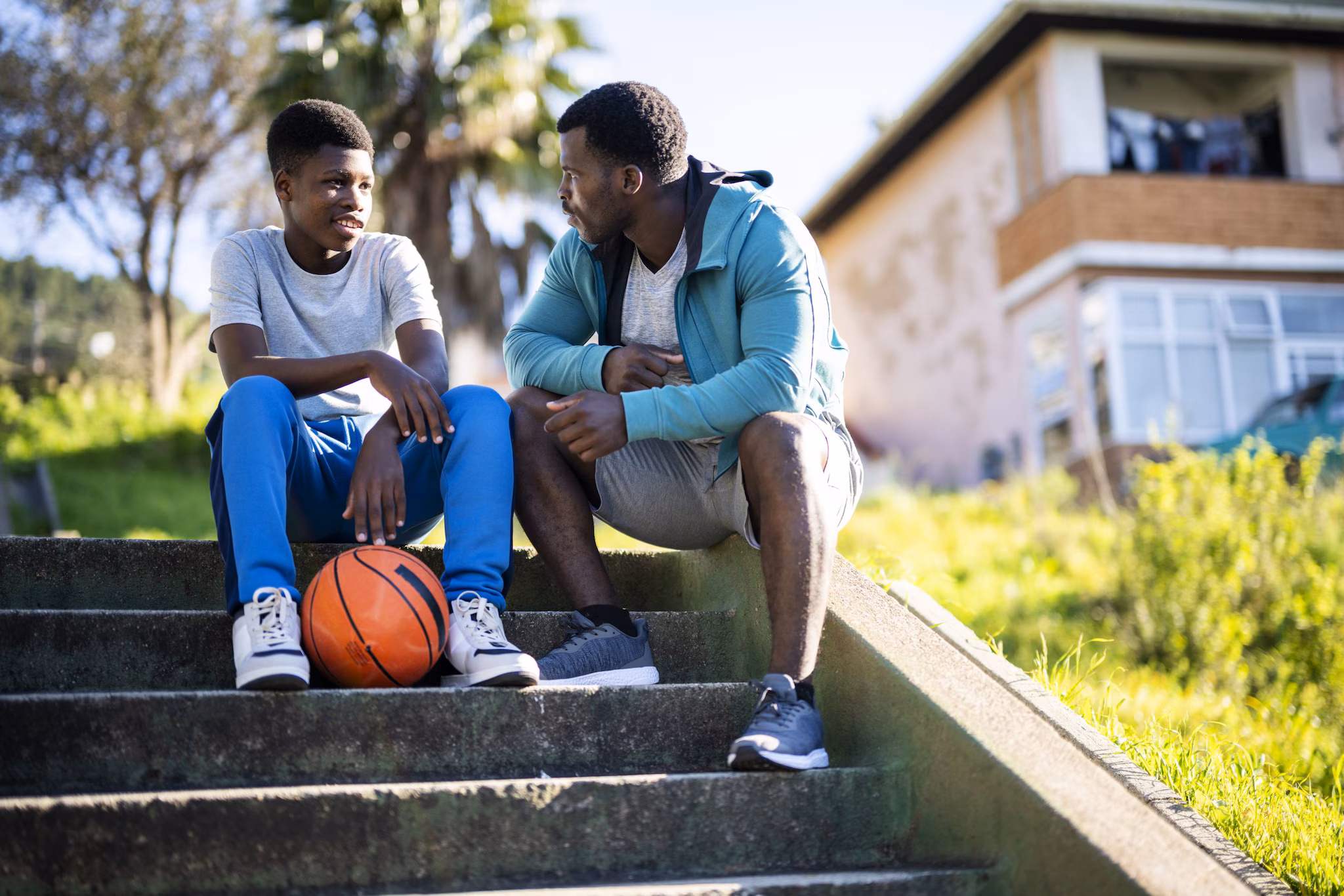 — title missing — A man and a child are sitting and talking on steps outside a housing area. A basketball lies at the feet of the young boy.