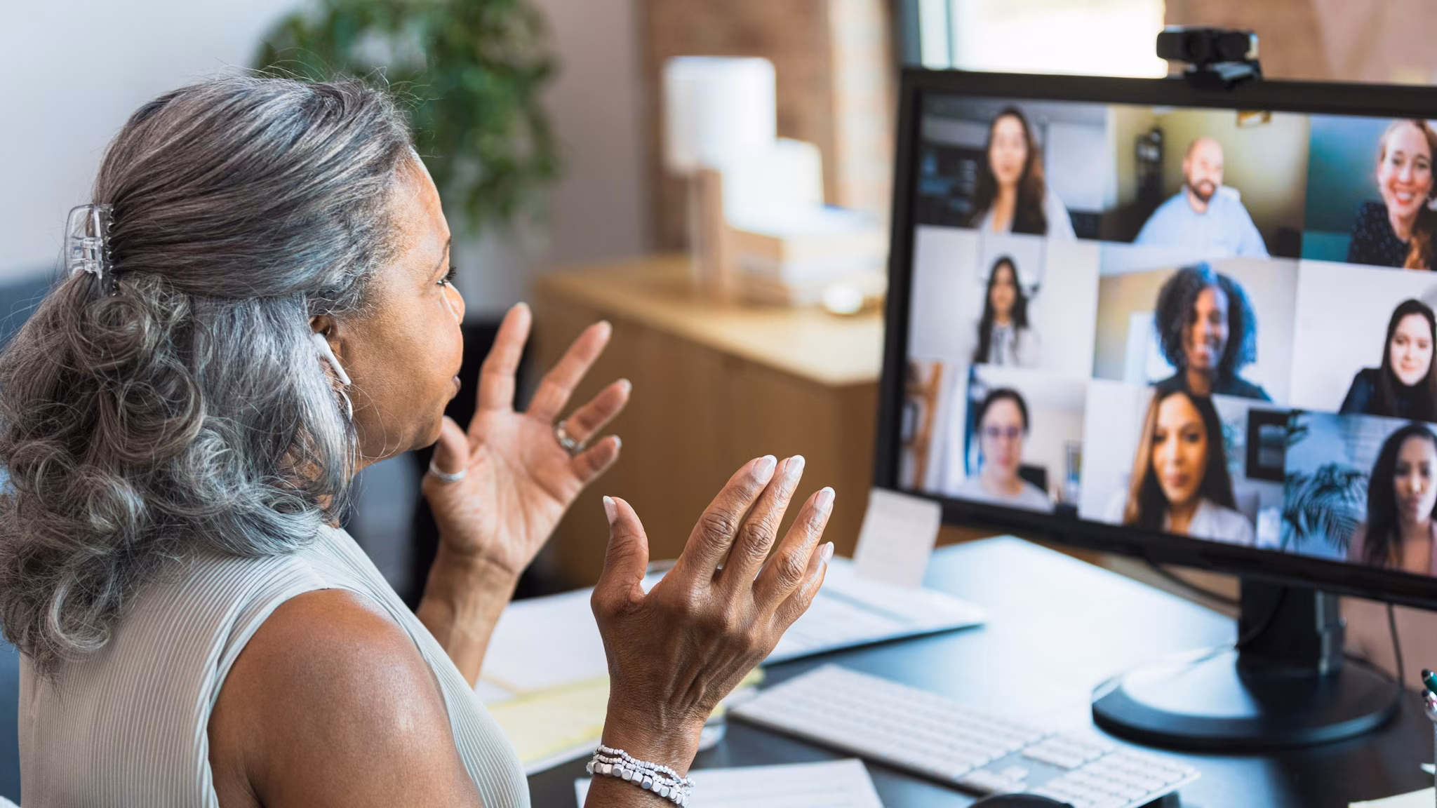 A woman sits in front of a computer and gives a virtual presentation to peers viewing on the screen.