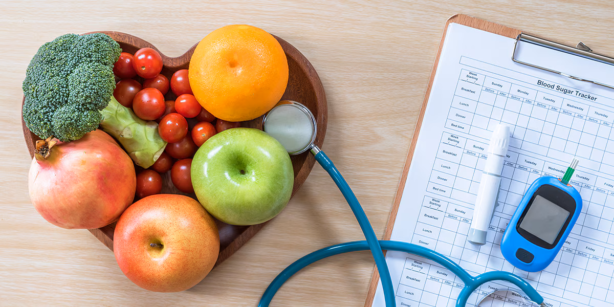 Diabetes monitor, Cholesterol diet and healthy food eating A heart shaped-bowl with colorful fruits and vegetables sits on a table with a stethoscope, glucose monitor, and clipboard.