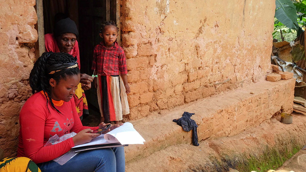 About FW image Community health worker sitting outside home, writing in notebook with woman and child looking on.