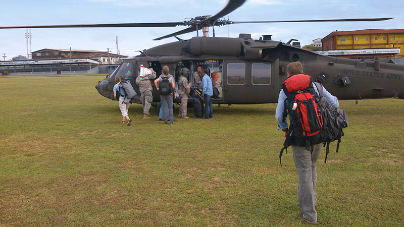Helicopter People boarding a U.S. Army helicopter on a grassy field, carrying backpacks and gear, ready for departure.