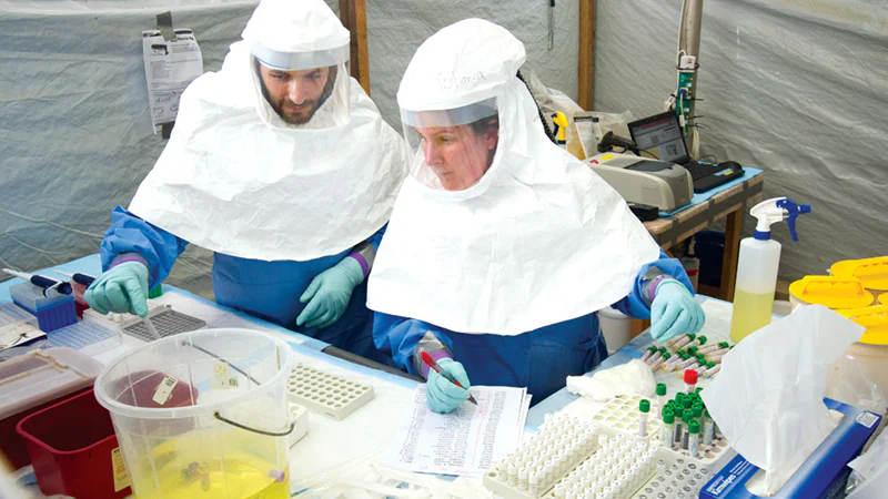 Lab Headgear Two scientists in protective suits working in a lab, handling samples and checking data.