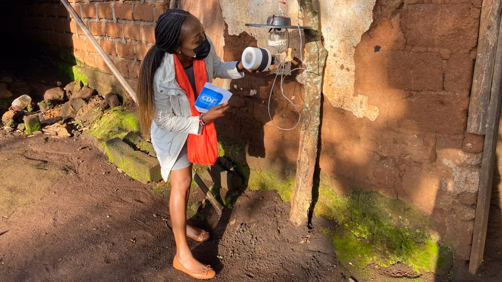 Cameroon_MosquitoLightTrap_2021 A woman examines a mosquito trap attached to a light outdoors.