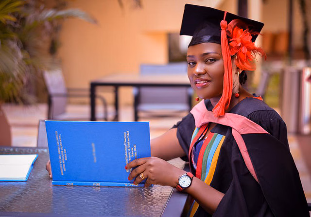 WFETP South Africa A woman wearing a black cap and gown with red accents is smiling while holding a blue book.