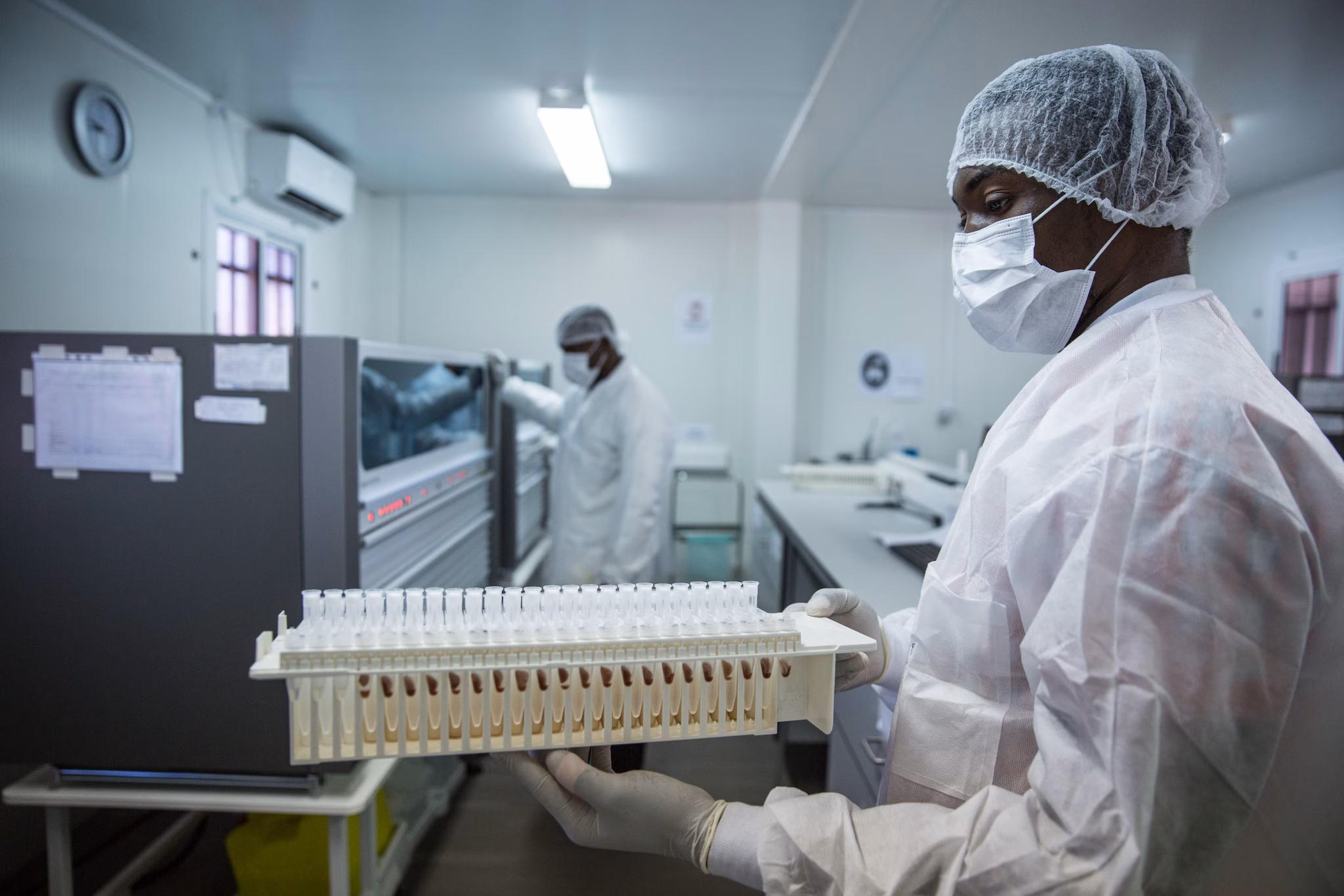 A Black man wearing white lab gear, blue gloves, a mask, and hair protection stands in a laboratory.