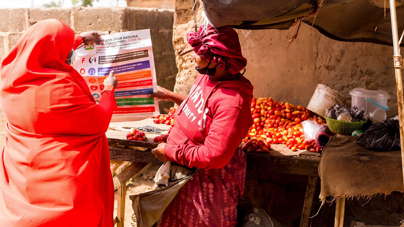 — title missing — An individual in bright orange holds up an informational poster. Another woman wearing red is looking at the poster as they stand outside.
