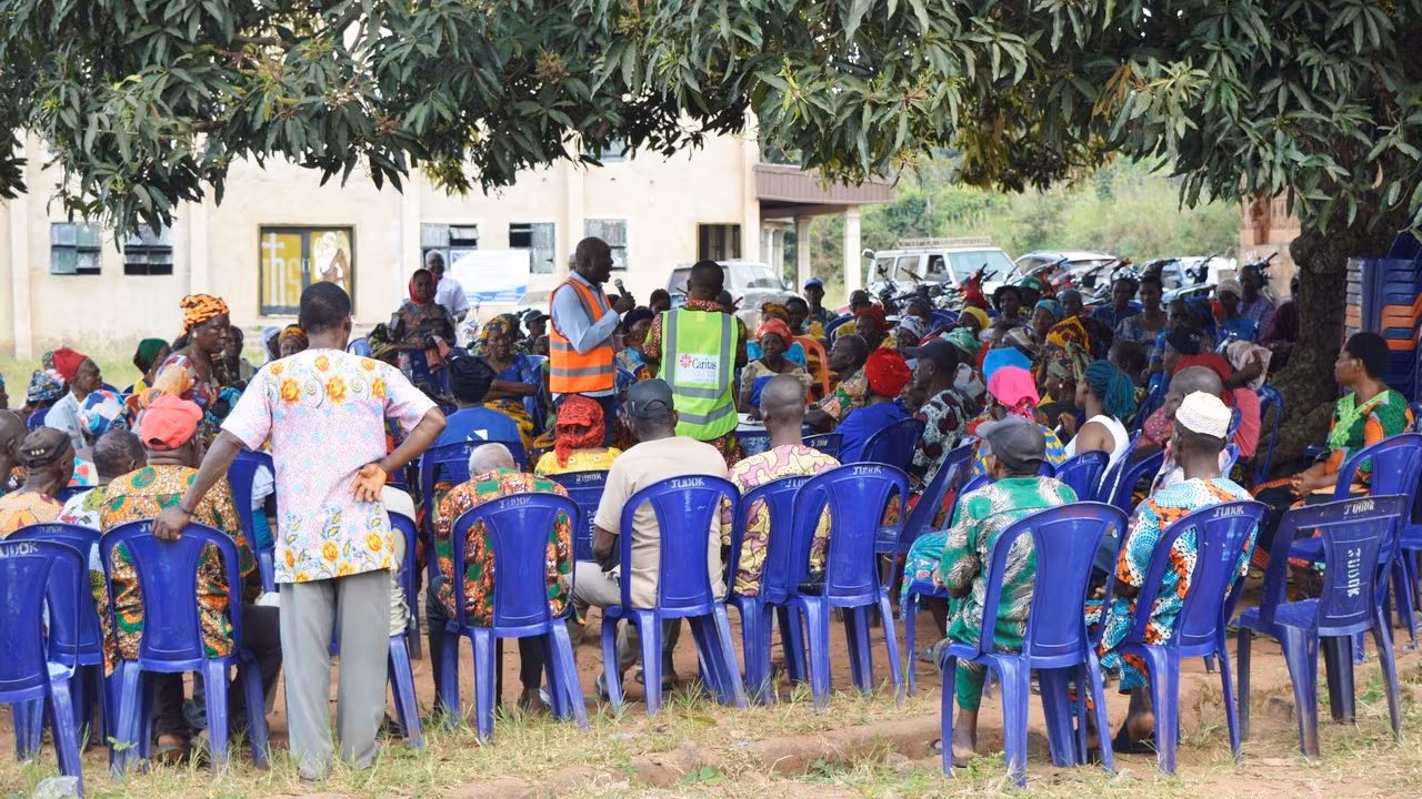 — title missing — A large group of individuals sit outside in chairs. A man with a microphone stands in the middle to share a message about COVID-19.