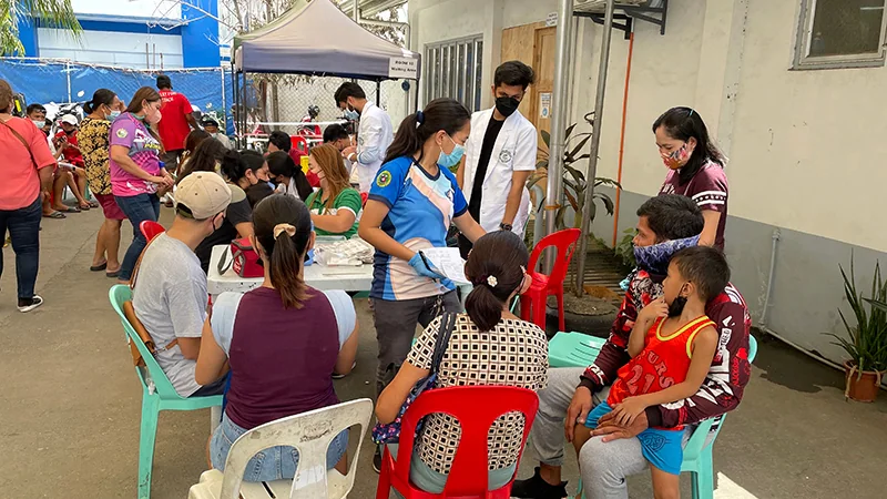 Philippines_HIVHealthWorkers_FieldPhoto Many people with face masks covering their nose and mouth are standing or sitting outdoors near a table and tent.