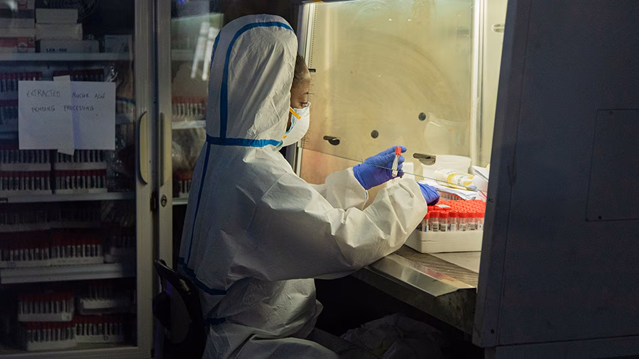 — title missing — A lab technician in personal protective equipment sits and works with testing supplies.