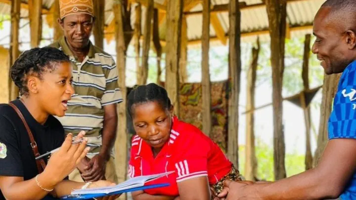 -- title missing -- A local community mobilizer in a black shirt sits with three other people, one in a brown stripe shirt, one in a red jersey, and one in a blue shirt, as she talks with them to collect data.