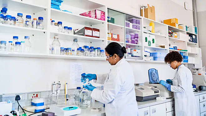 Two people in white coats and blue latex gloves work at a lab bench.