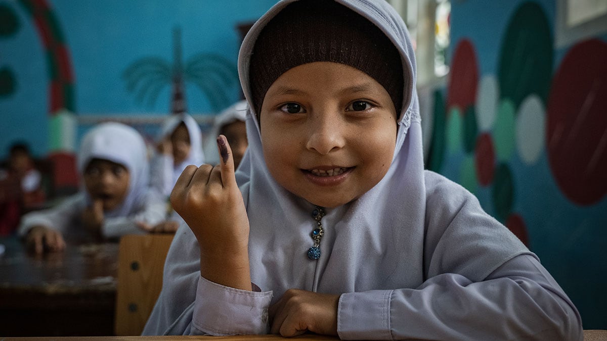 Young Indonesian girl holds up her pinky finger, marked to show she received a polio vaccine. A young girl sitting at a school desk smiles while she holds up her pinky finger, which has been marked to show she received a polio vaccine.