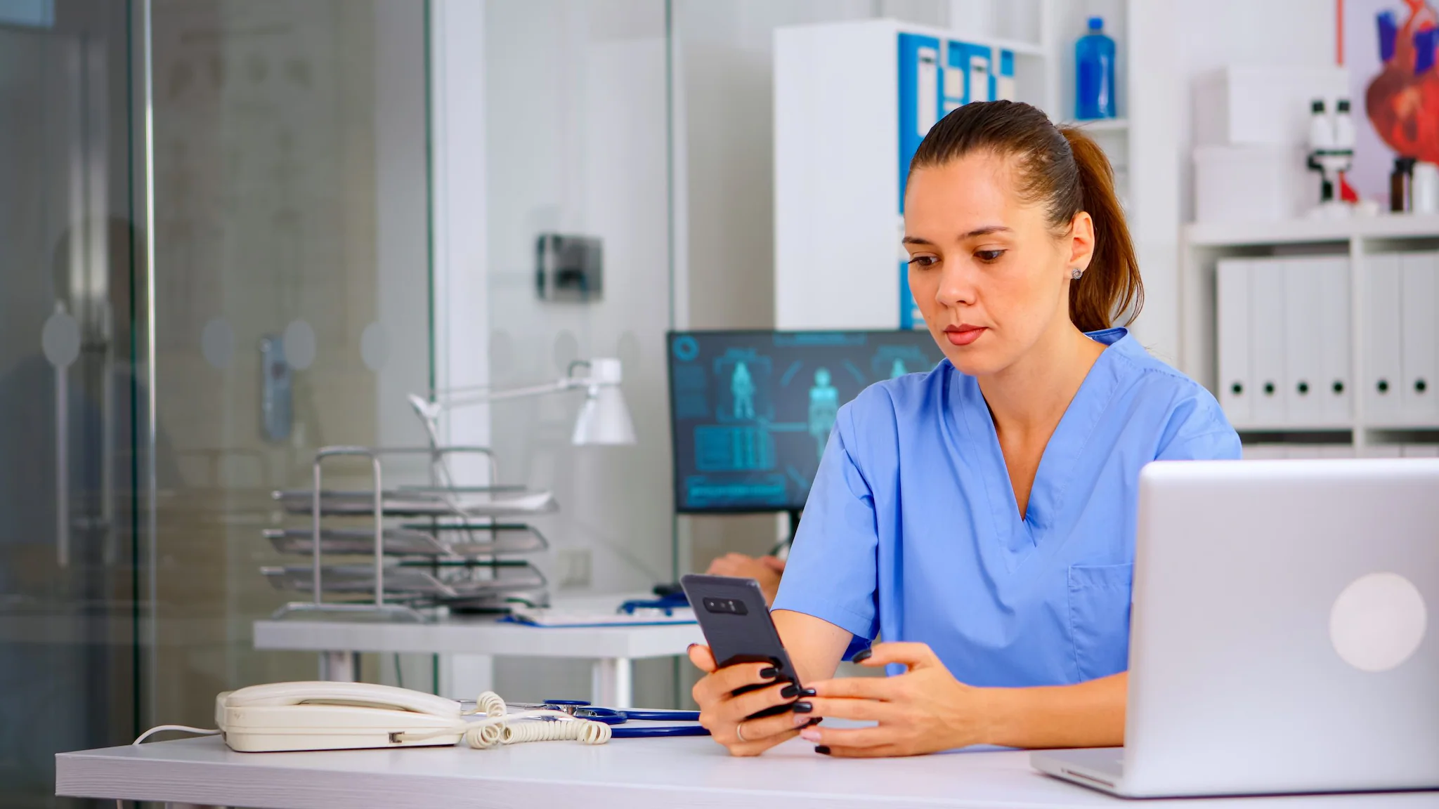 Using phone to calculate Medical professional sitting at a desk with a computer and a hand-held device.