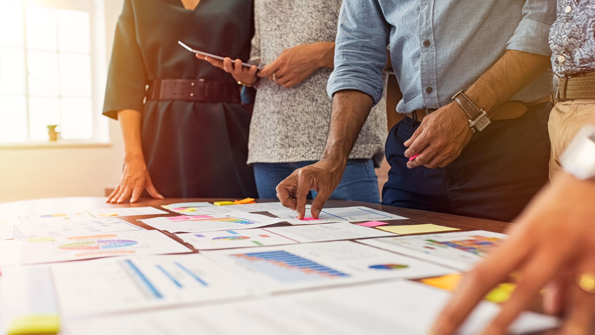 — title missing — A group of people standing around a table with pieces of paper with charts and graphs.
