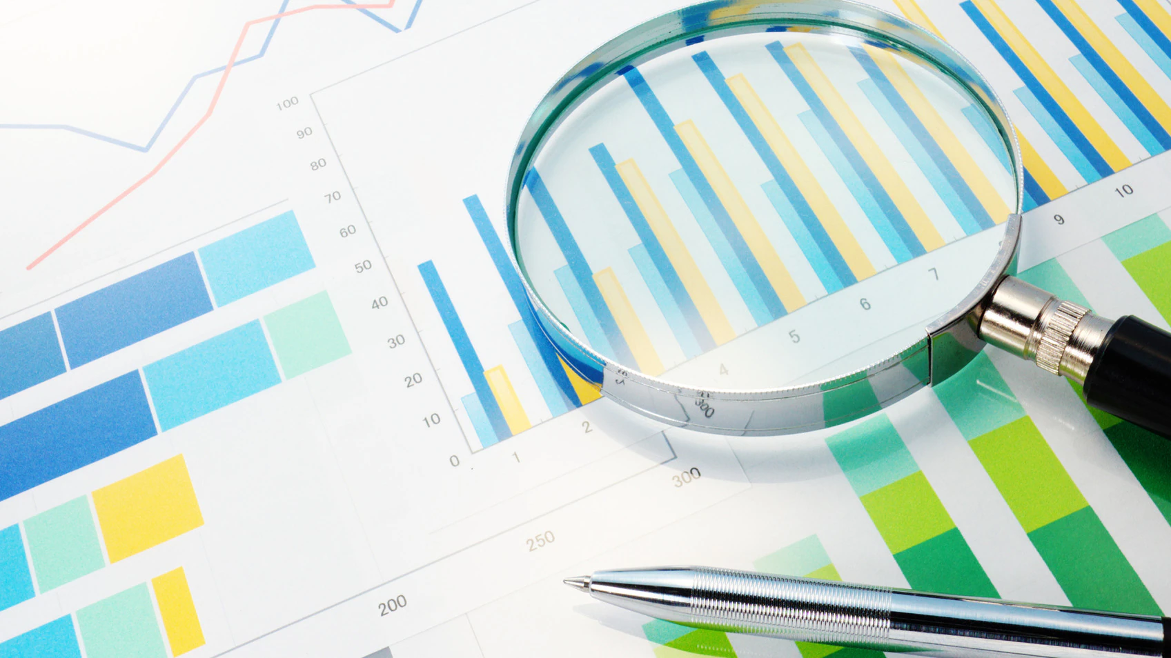 resized magnifying glass on paper Silver pen and magnifying glass on top of blue and green charts and figures.