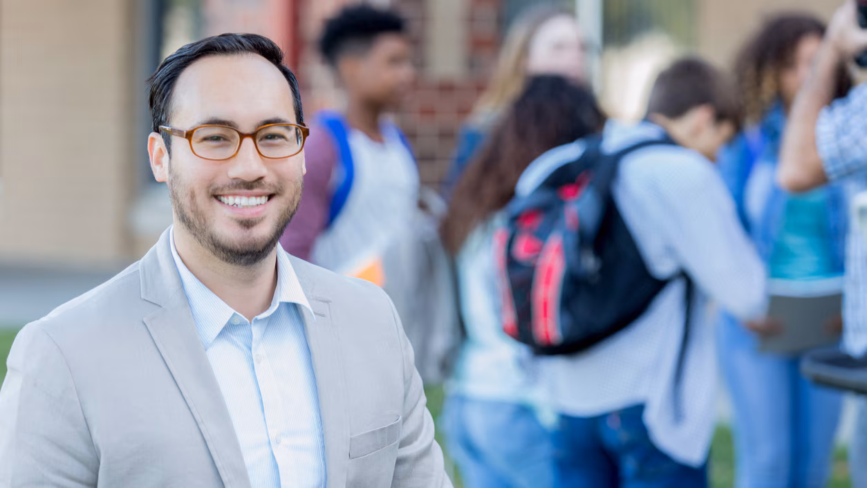 A high school teacher smiling with a group of students in the background.