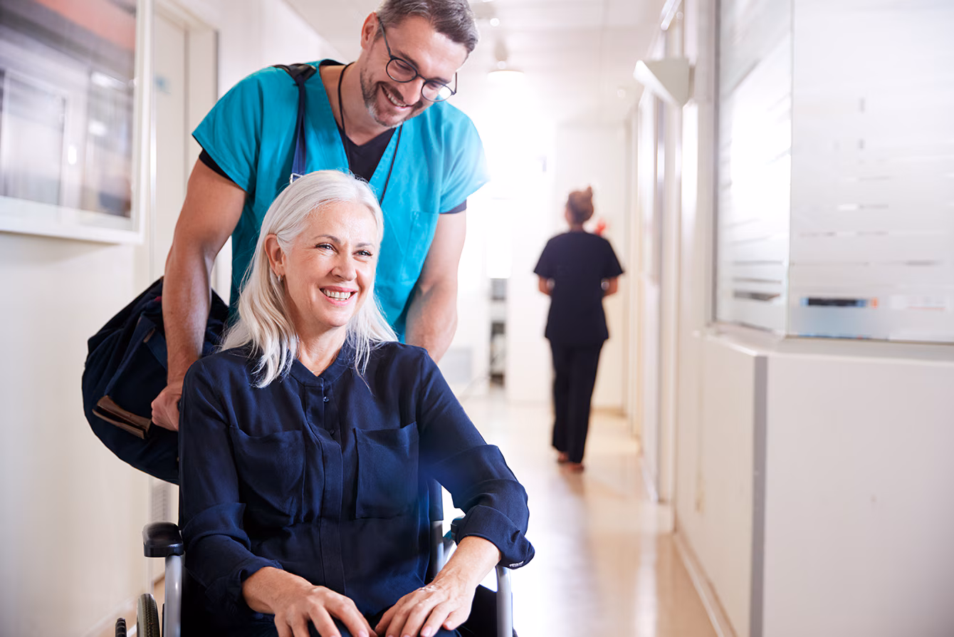 Patient-Discharge A smiling woman wearing a blue hoodie is sitting in a wheelchair being pushed through a hospital lobby to be discharged