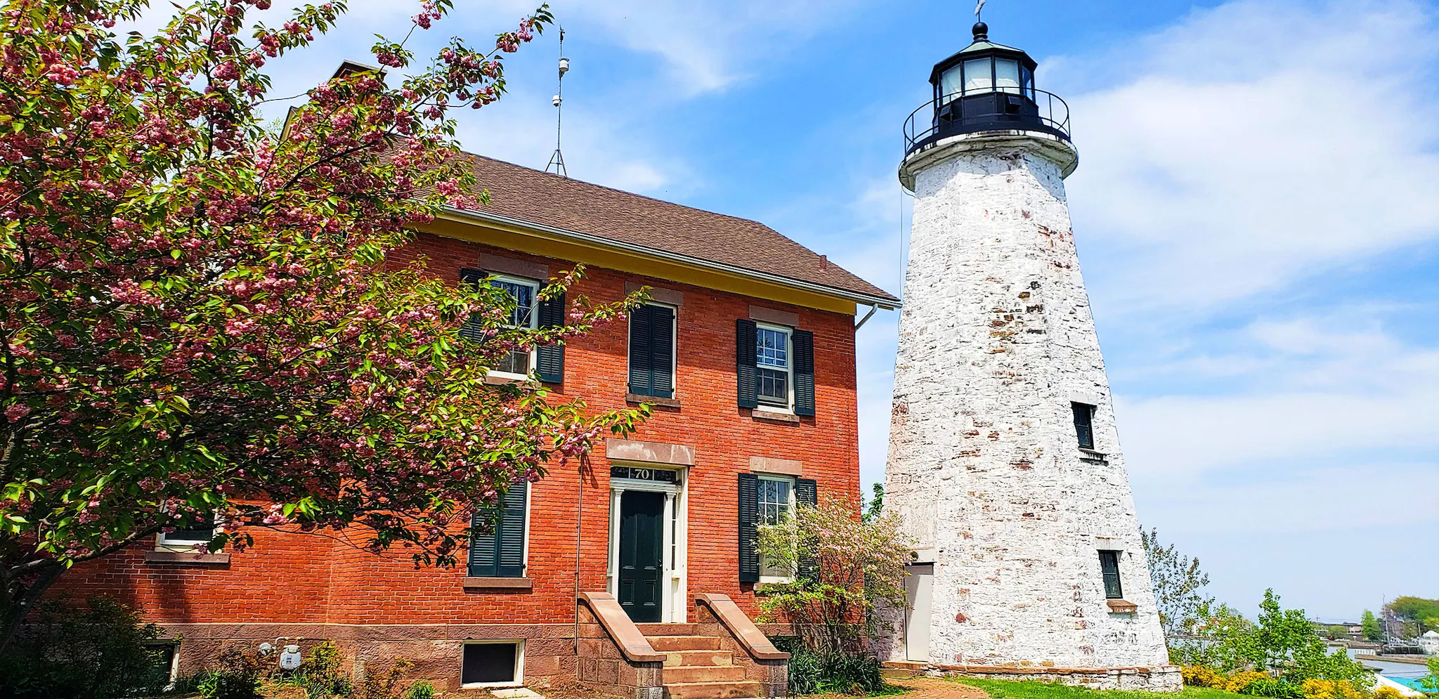 County of Genesee A red brick building with black shutters next to an old white lighthouse with a black top, surrounded by blooming trees and a clear blue sky.