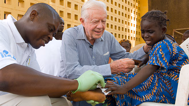 President Carter comforts a young girl as she receives medical care for Guinea worm disease.