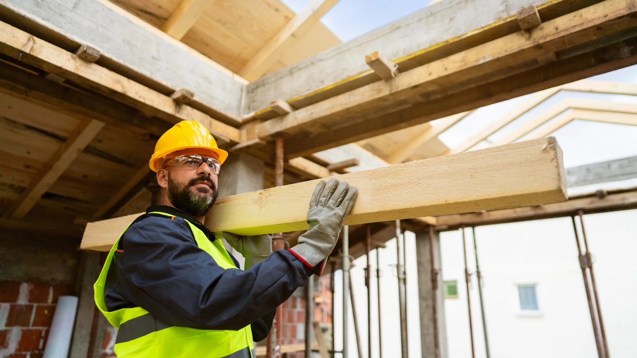 construction laborer Man with a beard wearing a hard hat, carrying lumber at a construction site.