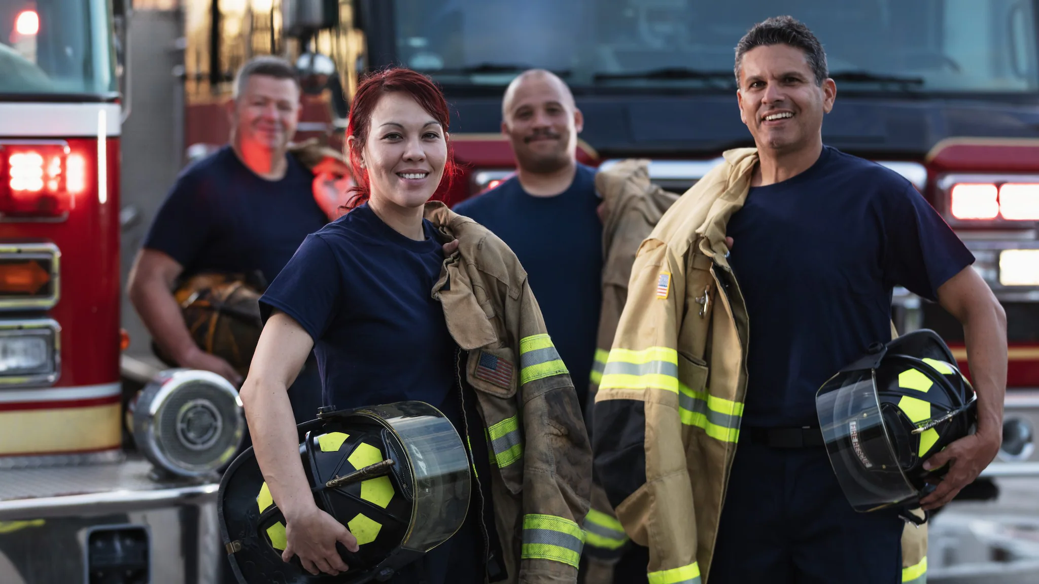 Group of FFs A multiracial group of four firefighters standing in front of fire engines holding PPE