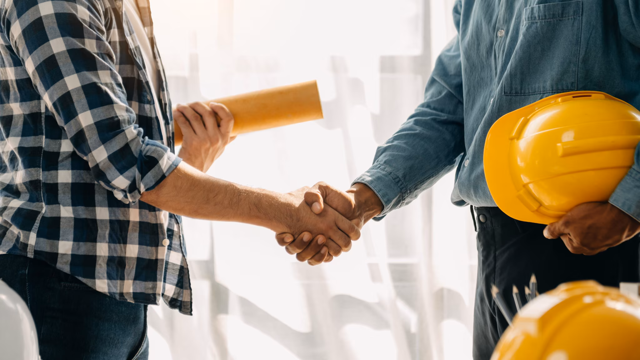 Construction team shake hands Two people shaking plans, one holds rolled up project plans and the other holds a safety helmet.