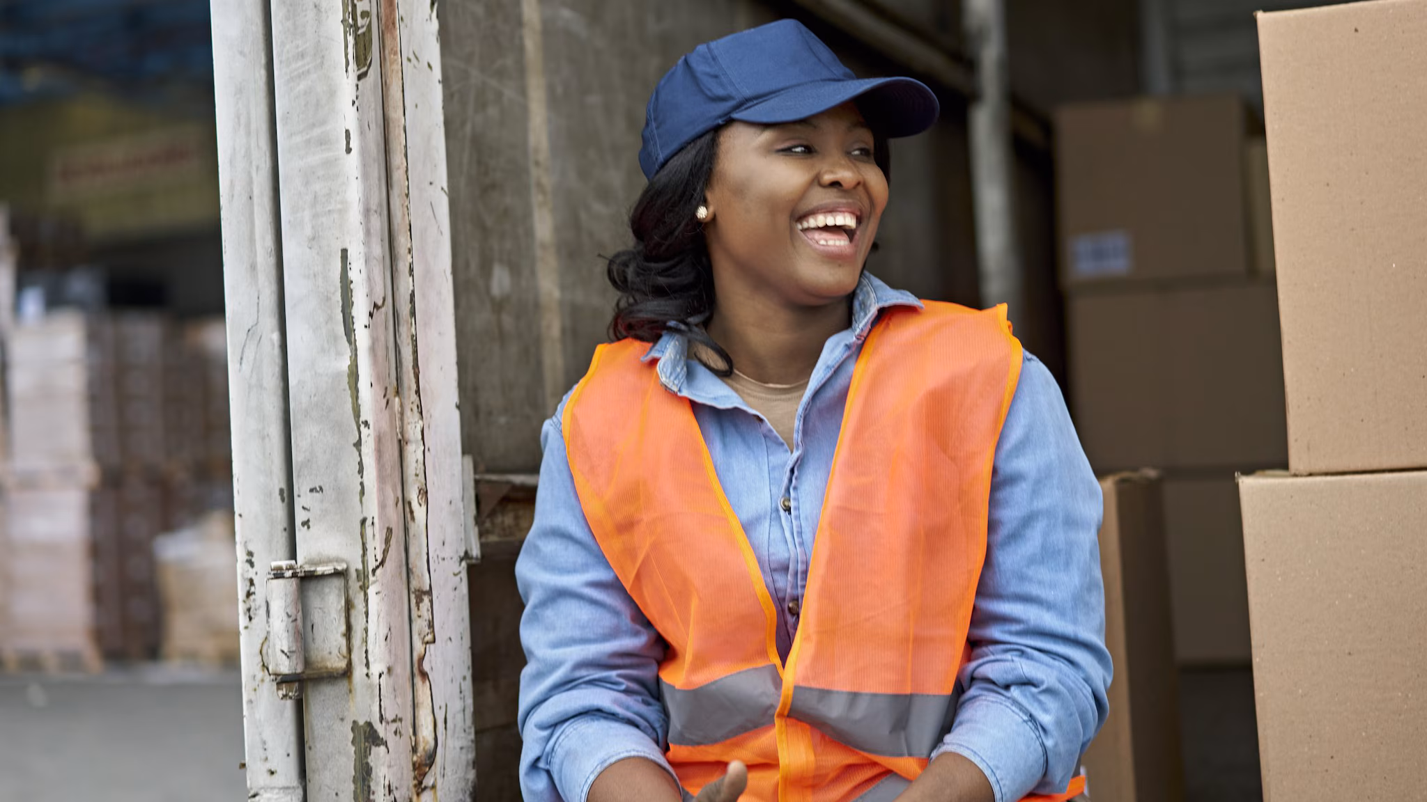 — title missing — woman wearing blue baseball hat and orange safety vest sitting on the back of a truck with boxes
