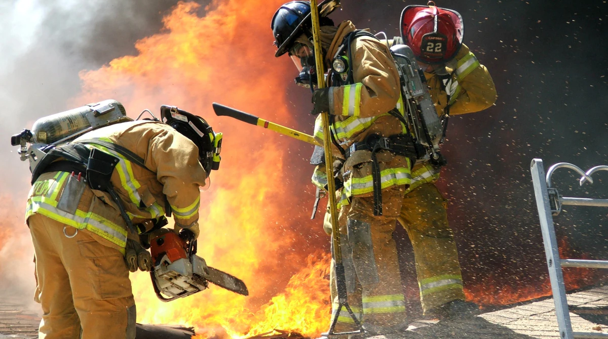 — title missing — Firefighters wearing PPE, carrying gear, near large flames.