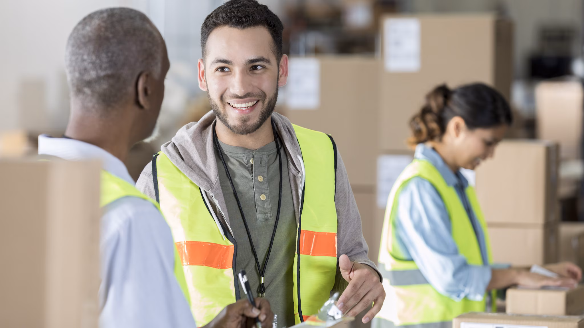 — title missing — Three warehouse workers wearing yellow safety vests. In the background, a worker affixes a label to a box. In the foreground, a worker is talking to his manager.