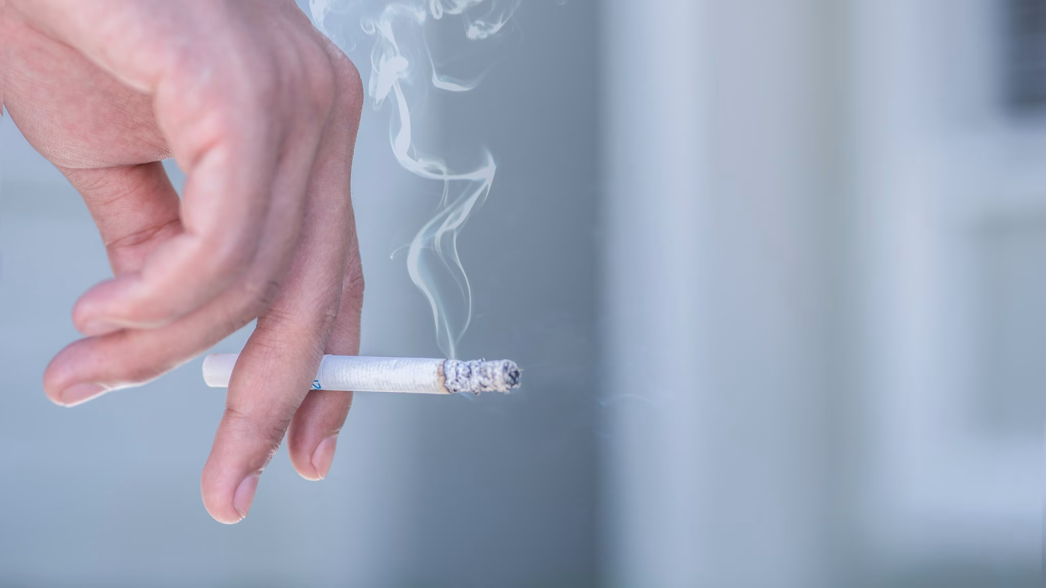 — title missing — A hand holding a lit cigarette with cigarette smoke flowing upward.