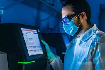 25137 A scientist wearing biosafety level 2 PPE gear including gloves, laboratory coat, goggles, and mask, using a sequencing instrument.