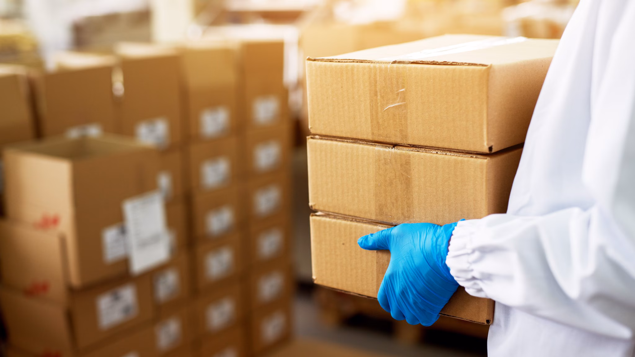 Person wearing a lab coat and blue gloves carrying cardboard boxes.
