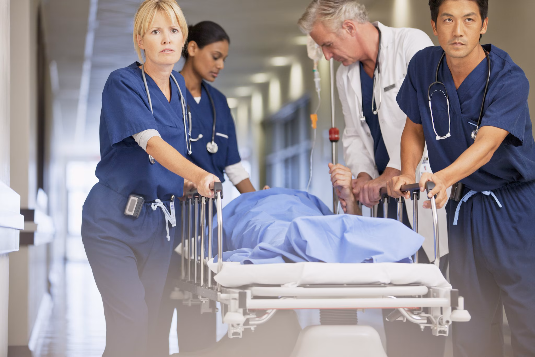 A four-person healthcare team rolling a patient draped in blue on a gurney through a hospital hallway