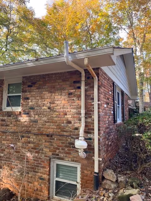 Corner view of a brick home with a pipe coming out of the side wall and up over the gutter to release radon into the air above the home.