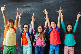 Happy Children Students in front of a chalk board smiling and happy with their arms raised over their heads.