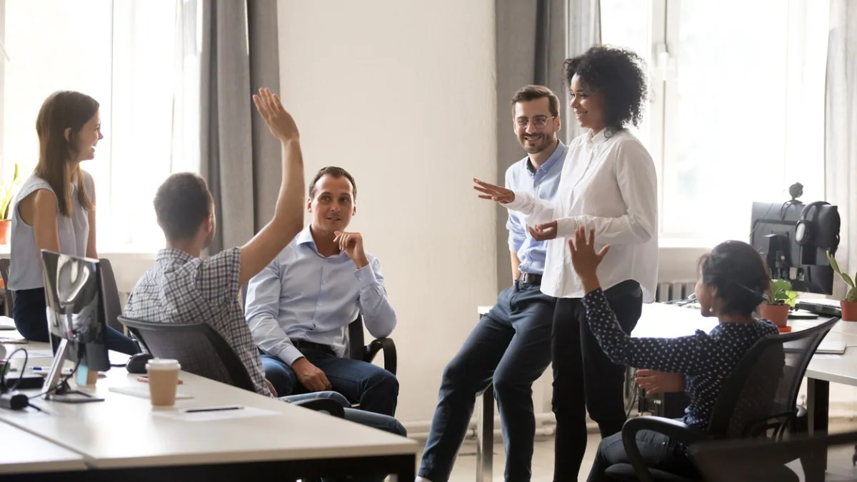 Coworkers_sharing_ideas_1200x675px Five coworkers talking and sharing ideas at work. One person is raising their hand.