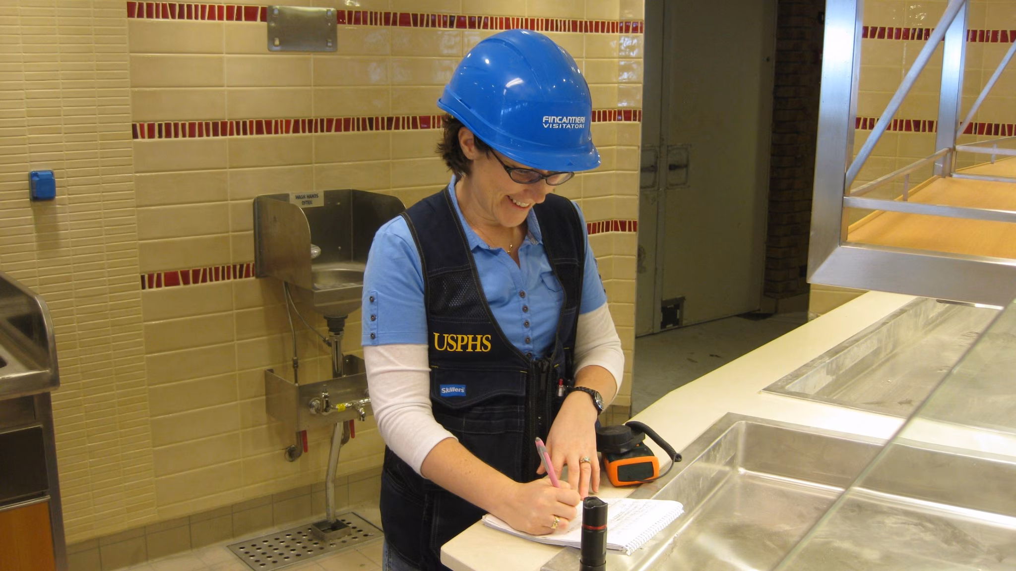 Public health officer wearing a hard hat taking notes on a clipboard inside a galley.